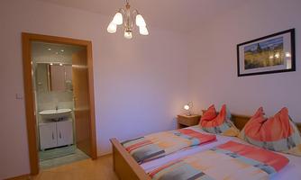 A cozy bedroom with a double bed and colorful cushions. A bathroom with modern facilities is visible in the background.