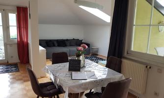 A bright, modern living room with a dining table and chairs. In the background, there is a comfortable sofa area with cushions.