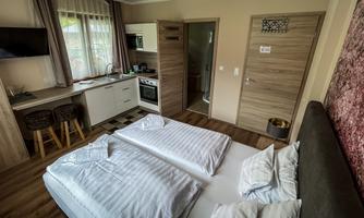 A modern room with two single beds, a kitchenette, and a small dining area. Large windows provide plenty of natural light.