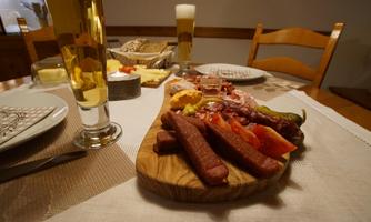 A wooden board with various types of sausages, fresh vegetables, and mustard. In the background, there are glasses of beer and a set table.