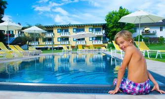 A boy is sitting at the edge of a swimming pool and smiling. In the background, there are loungers and sunny residential buildings.