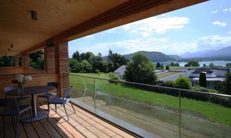 A balcony with a table and chairs offers a beautiful view of a green landscape and mountains. The sky is clear and sunny.