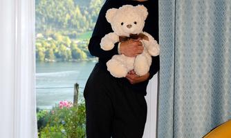 A man stands by a window holding a teddy bear. In the background, green hills and a lake can be seen.