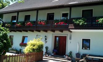 A cozy house with a balcony decorated with red flowers. In front of the house, there is a small fence and there are green plants.