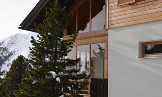 A modern house with wooden cladding and large windows. Surrounded by snow-covered mountains and fir trees.