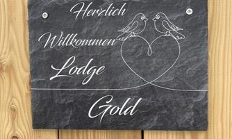 A warm welcome sign made of slate for the Gold Lodge, decorated with two birds and a heart. It is attached to a wooden wall.