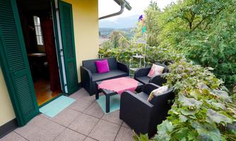 A cozy terrace with modern furniture and colorful cushions. In the background, there are green plants and a beautiful view.