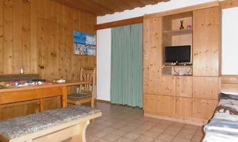 A cozy living space with wooden paneling and a large table. On the right side, there is a shelf with a TV and a couch.