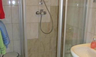 A small shower cabin with glass walls and a hand shower. Next to the shower is a sink, and some towels are hanging on the wall.