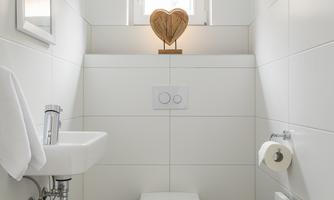 ‘The modern and functional toilet has a bright, friendly design. White wall tiles and dark flooring create clear contrasts and a neat overall look. A small window brings daylight into the room and is embellished with a decorative heart sculpture. The wall-mounted toilet, space-saving hand basin and practical accessories such as a mirror, waste bin and toilet brush blend harmoniously into the well-thought-out room concept.’