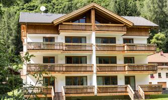 A modern building in the mountains, surrounded by green trees. The lawn in front of the house is well-kept and inviting.