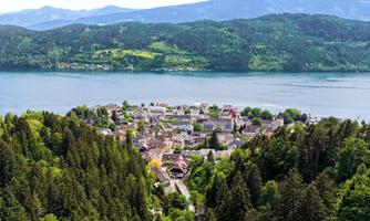 A picturesque village on the shore of a lake, surrounded by green forests and mountains. The sky is clear and the landscape is calm and inviting.