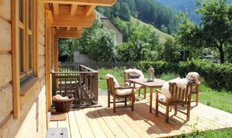 A cozy terrace with wooden furniture and soft blankets. In the background, green meadows and mountains can be seen.