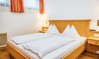 A cozy bedroom with a wooden bed and two white pillows. The walls are bright and the window coverings are in warm colors.