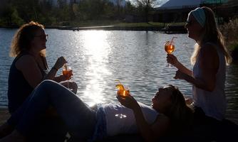 Three women are enjoying the sunset by the water, holding cocktails in their hands. The atmosphere is relaxed and friendly.