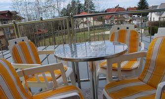 A cozy balcony with a round table and chairs in yellow and white. In the background, green landscapes and houses can be seen.