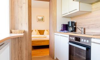A modern kitchen with wood and white cabinets. In the background, a bedroom with a cozy bed can be seen.