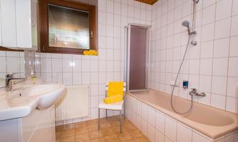 A modern bathroom with a bathtub, shower, and sink. The walls are tiled in white, and there are yellow towels on a chair.