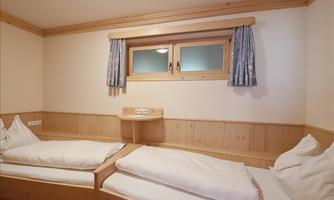 A simple bedroom with two single beds and light wooden paneling. The room has a window with curtains and a cozy atmosphere.