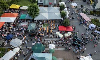 Ein belebter Markt mit bunten Ständen und vielen Menschen. Die Veranstaltung findet unter Zelten und Sonnenschirmen statt.
