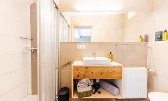 A modern bathroom with a sink on wooden furniture. The shower is separated by a glass wall and there are some toiletries on the shelf.
