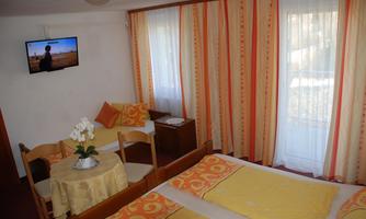 A simple room with two beds, yellow bedding, and a small dining table. A television hangs on the wall and the windows are adorned with white curtains.