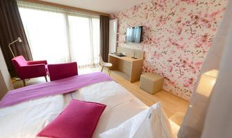 A modern hotel room with a double bed and a pink decorative blanket. Large windows provide plenty of daylight, and the walls are decorated with a floral pattern.