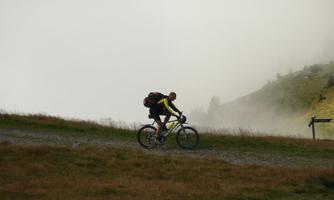 A cyclist rides on a narrow path through a misty landscape. The surroundings are green with gentle hills.