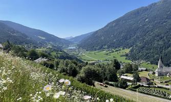 A picturesque valley with green meadows and gentle hills. In the foreground, white flowers are blooming and a small village with a church is visible.