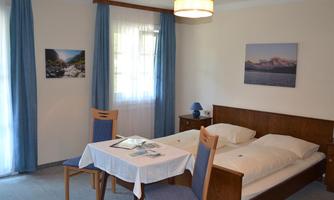 A bright room with a double bed and a table. The windows are adorned with blue curtains, and pictures decorate the walls.