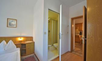 A simple room with a bed and a small table. In the background, a bathroom and a kitchen are visible.