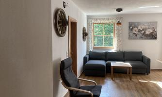 A bright living space with a gray sofa and a wooden chair. A window lets in plenty of daylight, and a wall decoration with flowers is visible.