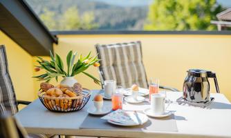 A cozy breakfast table on a balcony with fresh rolls, coffee, and fresh flowers. In the background, a green landscape can be seen.