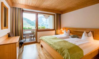 A cozy hotel room with a double bed and wooden decor. Through the window, there is a view of the mountains and a terrace.
