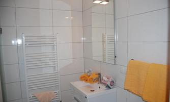 A modern bathroom with white tiles and a large mirror. Yellow towels are on the sink and a chair.
