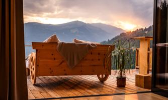A cozy wooden lounge chair is on a porch with a view of the mountains. In the background, a beautiful sunset can be seen.