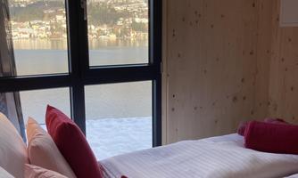 A cozy bedroom with a view of the lake and the mountains. The beds are decorated with red pillows and there are large windows that let in plenty of light.