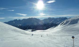 A snow-covered mountain landscape with skiers. The sky is clear and the sun shines brightly.