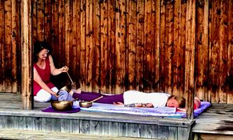A woman is lying relaxed on a mat while another woman is working with sound bowls. The background consists of wood paneling that creates a calming atmosphere.
