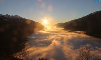 An impressive view of a valley above a blanket of clouds at sunrise. The mountains in the background are partially covered with snow.