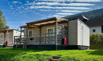 A modern holiday home with a porch stands in a green meadow. The sky is clear with a few clouds.