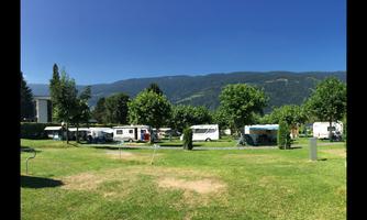 A campsite with several caravans and green trees. In the background, gentle mountains and a bright blue sky can be seen.