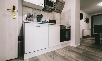 A modern kitchen with white cabinets and wooden flooring. It is well-equipped with a stove, a sink, and kitchen appliances.