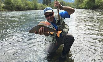 A fisherman stands in the water and proudly holds a caught fish. In the background, green trees and a picturesque landscape can be seen.