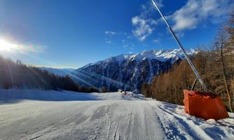 A snowy ski slope with sunlight illuminating the mountains. In the background, majestic mountain peaks rise under a clear blue sky.