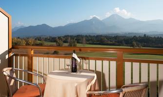 A cozy terrace with a table and two chairs, overlooking a beautiful mountain landscape. On the table, there is a bottle of wine, while the sun shines over the mountains.