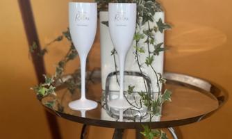 Two elegant glasses are standing on a round table. In the background, there are green plants and an orange wall.