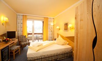 A cozy hotel room with a double bed, a television, and a small table. The large windows offer a view of the balcony and the beautiful scenery.