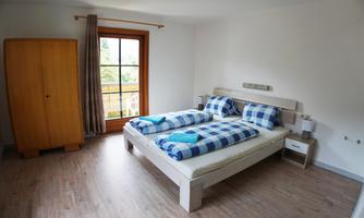 A bright bedroom with a double bed and checked blankets. A large window overlooking the outside and a wooden box complete the space.