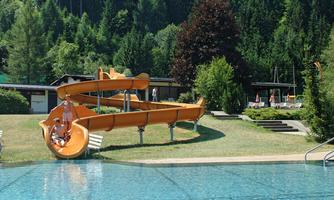 A swimming pool with a large, orange water slide. Surrounded by green trees and deck chairs in the background.